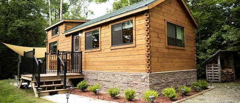 A wooden cabin with a stone foundation, a front porch, and a small shed in a wooded area.