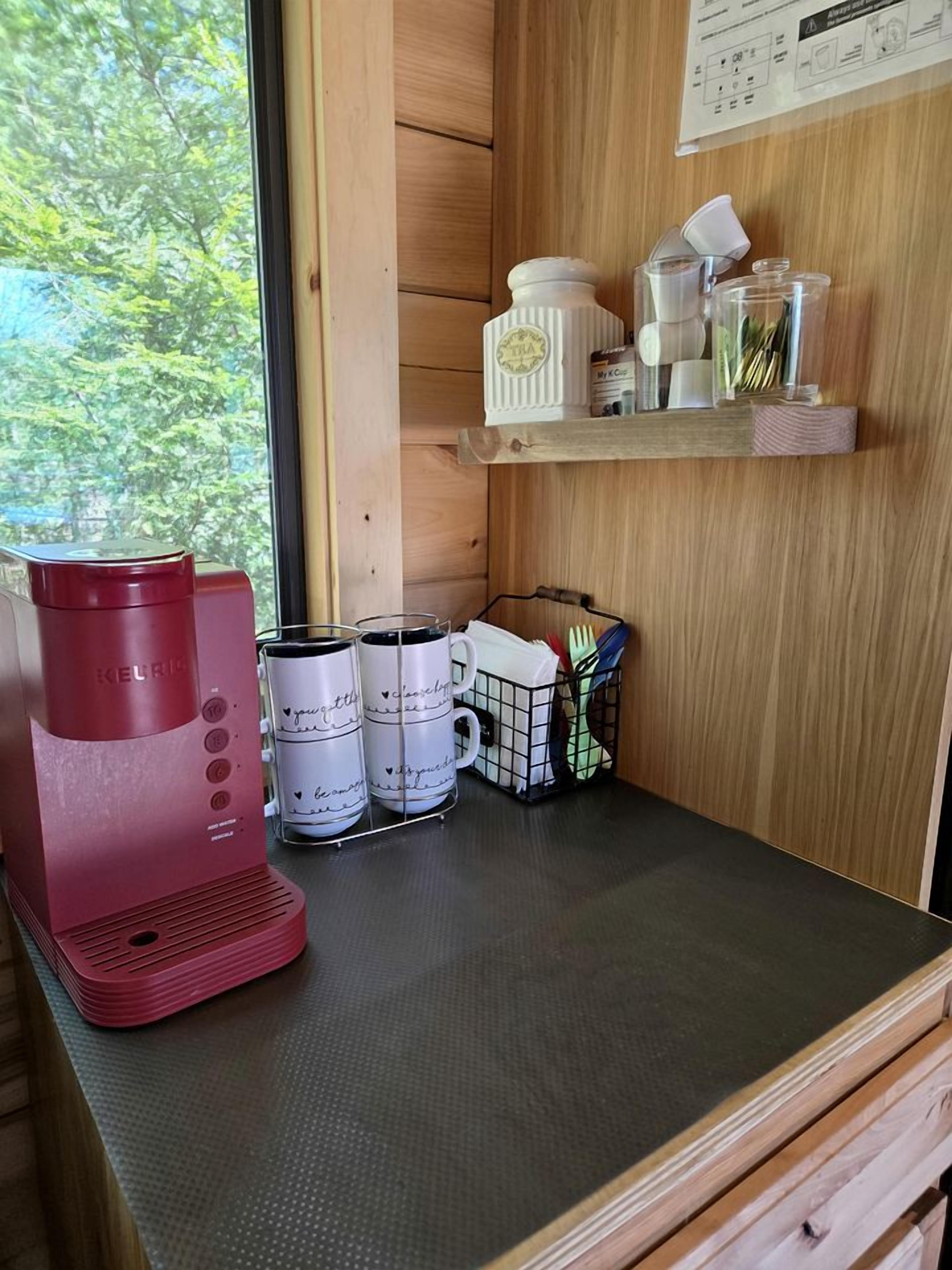 Une petite cuisine équipée avec une machine à café rouge, des tasses blanches ornées de motifs, une petite étagère contenant des ustensiles de cuisine et une fenêtre donnant sur des arbres.