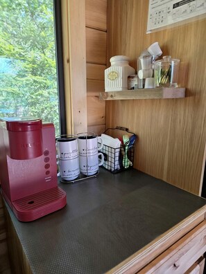 Eine Küchenzeile mit einer roten Kaffeemaschine, weißen Tassen mit Aufdruck, einer kleinen Ablage mit Küchenutensilien und einem Fenster mit Blick auf Bäume.