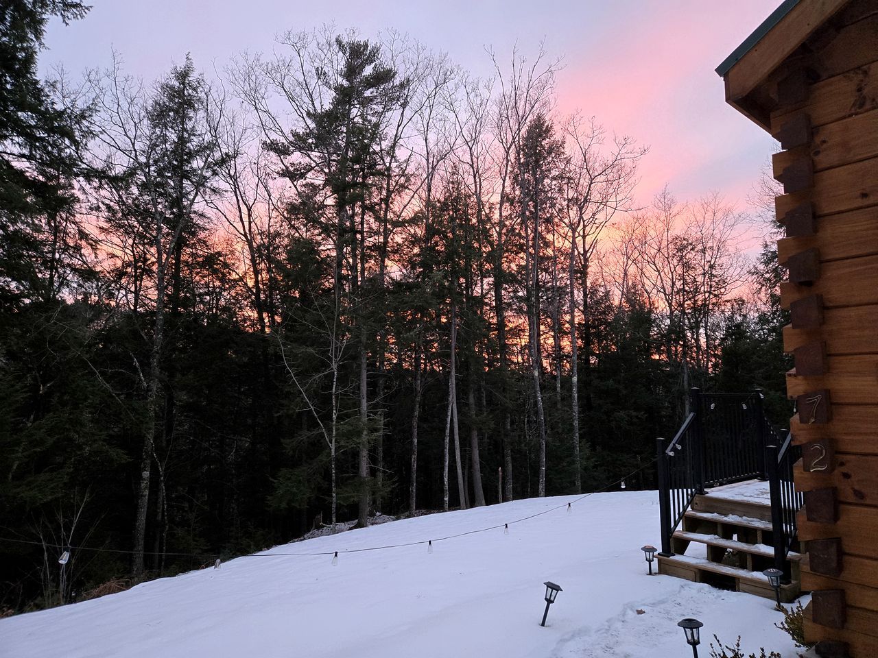 A wooden cabin with a staircase leading to a deck, surrounded by a snowy landscape and trees.