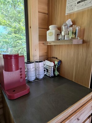 Eine Küchenzeile mit einer roten Kaffeemaschine, weißen Tassen mit Aufdruck, einer kleinen Ablage mit Küchenutensilien und einem Fenster mit Blick auf Bäume.