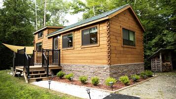 A wooden cabin with a stone foundation, a front porch, and a small shed in a wooded area.