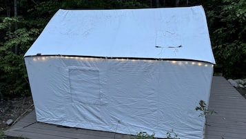 Una carpa blanca con techo plano y luces colgantes sobre un piso de madera, con un bosque de fondo.