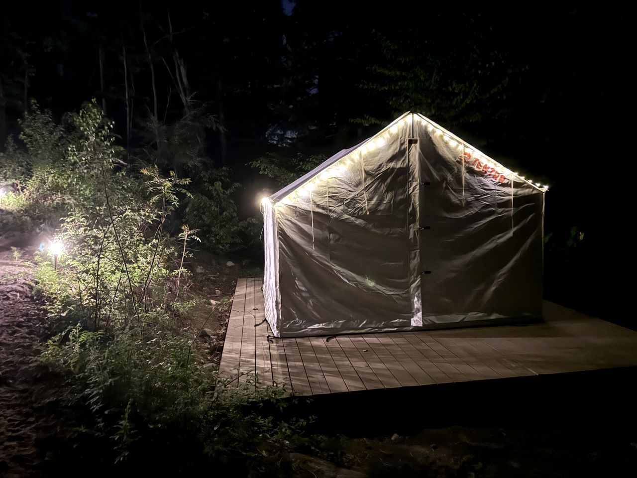 A tent-like structure with a roof and walls, illuminated by artificial light, set on a wooden platform surrounded by vegetation.
