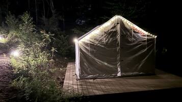 A tent-like structure with a roof and walls, illuminated by artificial light, set on a wooden platform surrounded by vegetation.