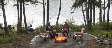 Une terrasse en bois avec un foyer, entourée de chaises, donnant sur un lac avec des arbres en arrière-plan.
