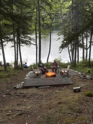 Une terrasse en bois avec un foyer, entourée de chaises, donnant sur un lac avec des arbres en arrière-plan.