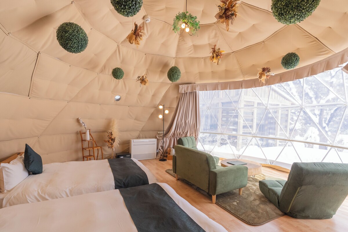 A cozy, well-lit tent-style room with two beds, a sofa, and a large window offering a view of a snowy landscape.