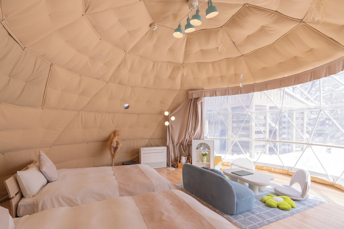 A cozy, well-lit tent-style room with a bed, seating area, and a window offering a view of the outside.