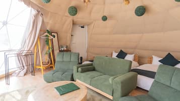 A tented accommodation with a green sofa, a round wooden table, and a bed with pillows.