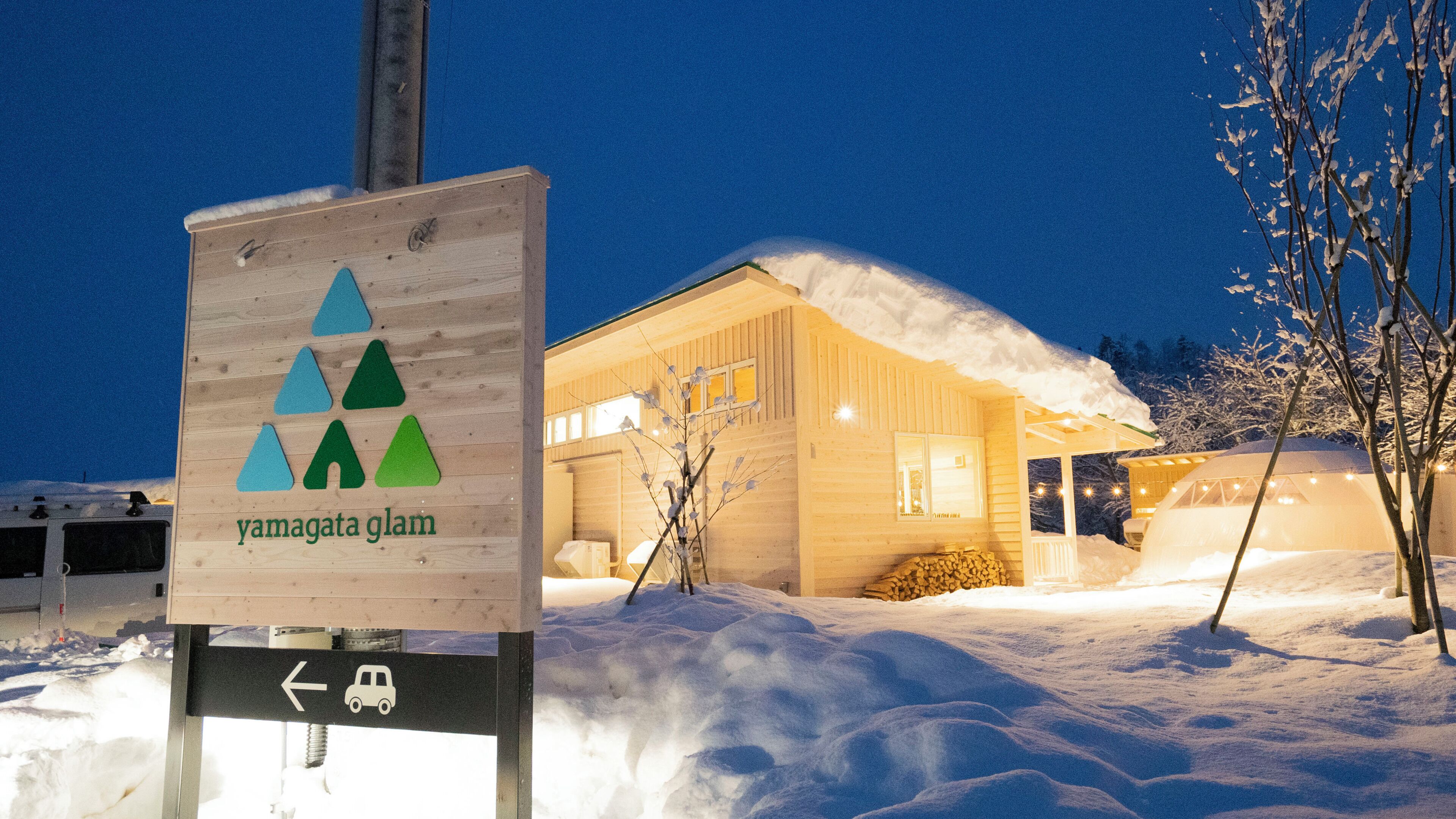 Une cabane en bois dont le toit est recouvert de neige, une enseigne ornée de symboles de montagne et un pictogramme de voiture indiquant la présence d’un parking à proximité.
