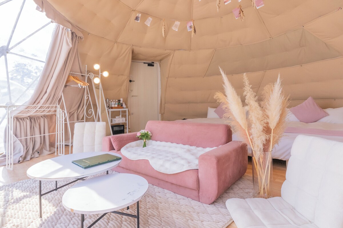 A tented accommodation with a pink sofa, white armchairs, a small table, and pampas grass decoration.