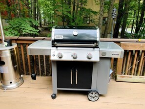 Een draagbare Weber Genesis II barbecue op een houten terras, met een metalen prullenbak en een houten omheining op de achtergrond.