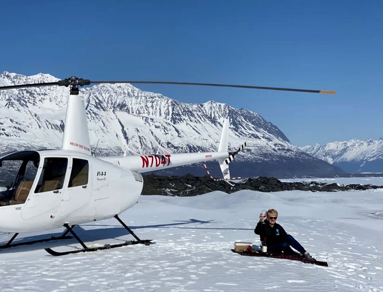 A helicopter with a person sitting on the snow beside it.
