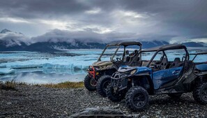 Three off-road vehicles parked on a rocky shore with a glacier and mountains in the background.