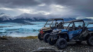 Three off-road vehicles parked on a rocky shore with a glacier and mountains in the background.