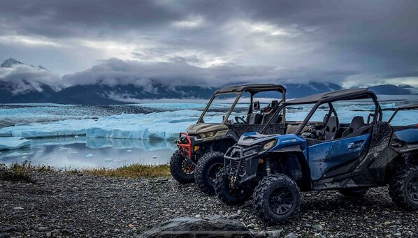 Tres vehículos todoterreno estacionados en una costa rocosa, con un glaciar y montañas al fondo.