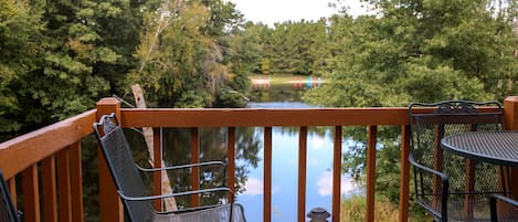 A wooden deck with metal chairs and a table overlooking a calm body of water and dense greenery.