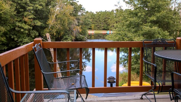 A wooden deck with metal chairs and a table overlooking a calm body of water and dense greenery. - Family-Friendly Cabin Rental on a Private Lake near Wisconsin Dells (Tomah)