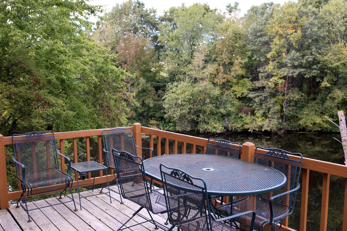 A wooden deck with a round table and chairs, surrounded by dense trees and a body of water.