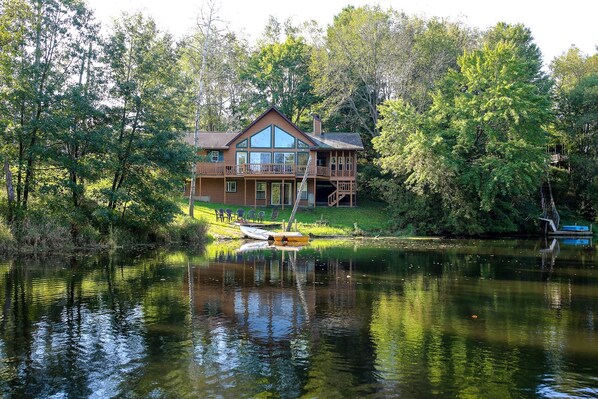A two-story log cabin with a balcony, situated by a calm lake surrounded by dense trees. - Family-Friendly Cabin Rental on a Private Lake near Wisconsin Dells (Tomah)