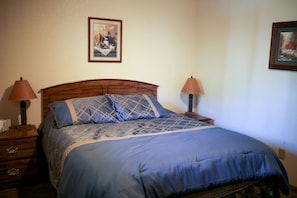 Ein Schlafzimmer mit einem hölzernen Kopfteil, einer blauen Bettdecke, zwei Nachttischlampen und einem gerahmten Bild an der Wand.