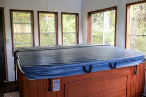 Ein Whirlpool mit blauer Abdeckung, Holzwänden und mehreren Fenstern mit Blick auf Bäume.