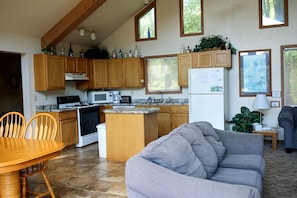 Eine Küche mit Holzschränken, einem Herd, einer Mikrowelle und einem Kühlschrank; ein Essbereich mit Tisch und Stühlen; und ein Wohnbereich mit einer Couch.