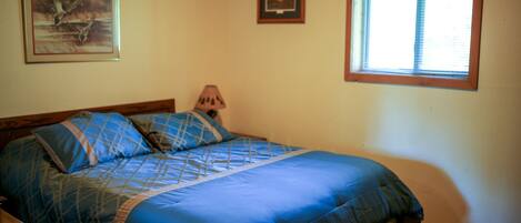 Un dormitorio con cama, mesita de noche, lámpara, ventana con cortinas y dos cuadros en la pared.