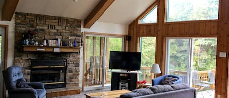 A cozy living room with a stone fireplace, a flat-screen TV, and a large window offering a view of the outdoors.