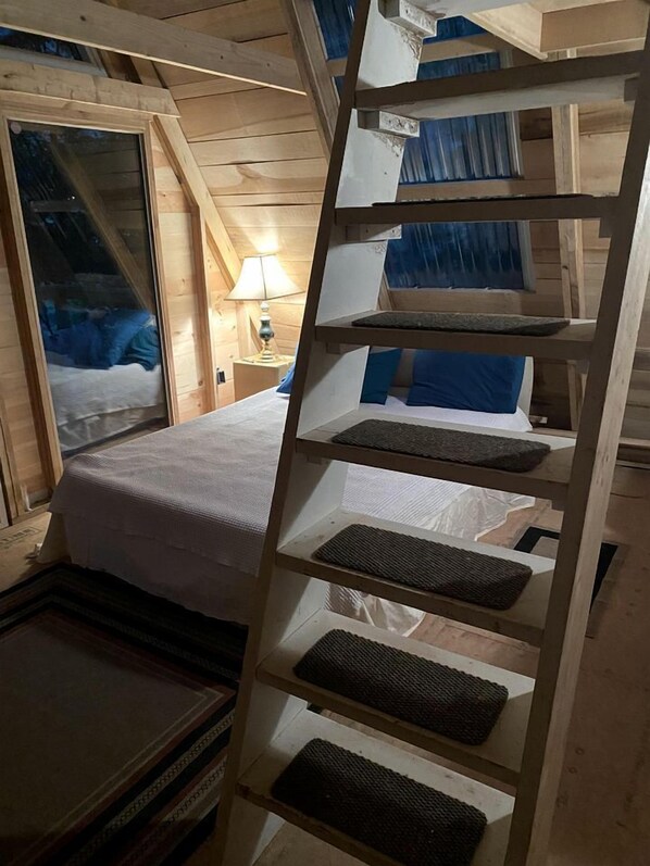 Interior de una cabaña de madera con una litera, una escalera y una ventana con cortinas.