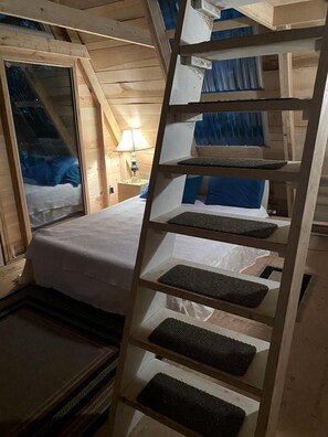 Interior de una cabaña de madera con una litera, una escalera y una ventana con cortinas.