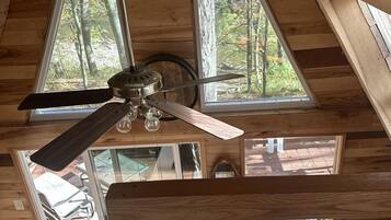 A wooden cabin interior with a ceiling fan, large windows, a guitar, and a sofa.