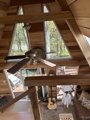 Interior de una cabaña de madera con ventilador de techo, ventanas amplias, una guitarra y un sofá.