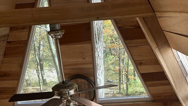 Interior de una cabaña de madera con ventilador de techo, ventanas amplias, una guitarra y un sofá.