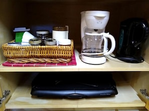 A kitchen shelf with a coffee maker, a basket of jars, and a Proctor Silox appliance.