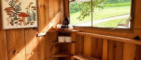 Interior d'una cabana de fusta amb finestra que dona a un paisatge verd, una estant amb espelmes i una il·lustració botànica emmarcada.