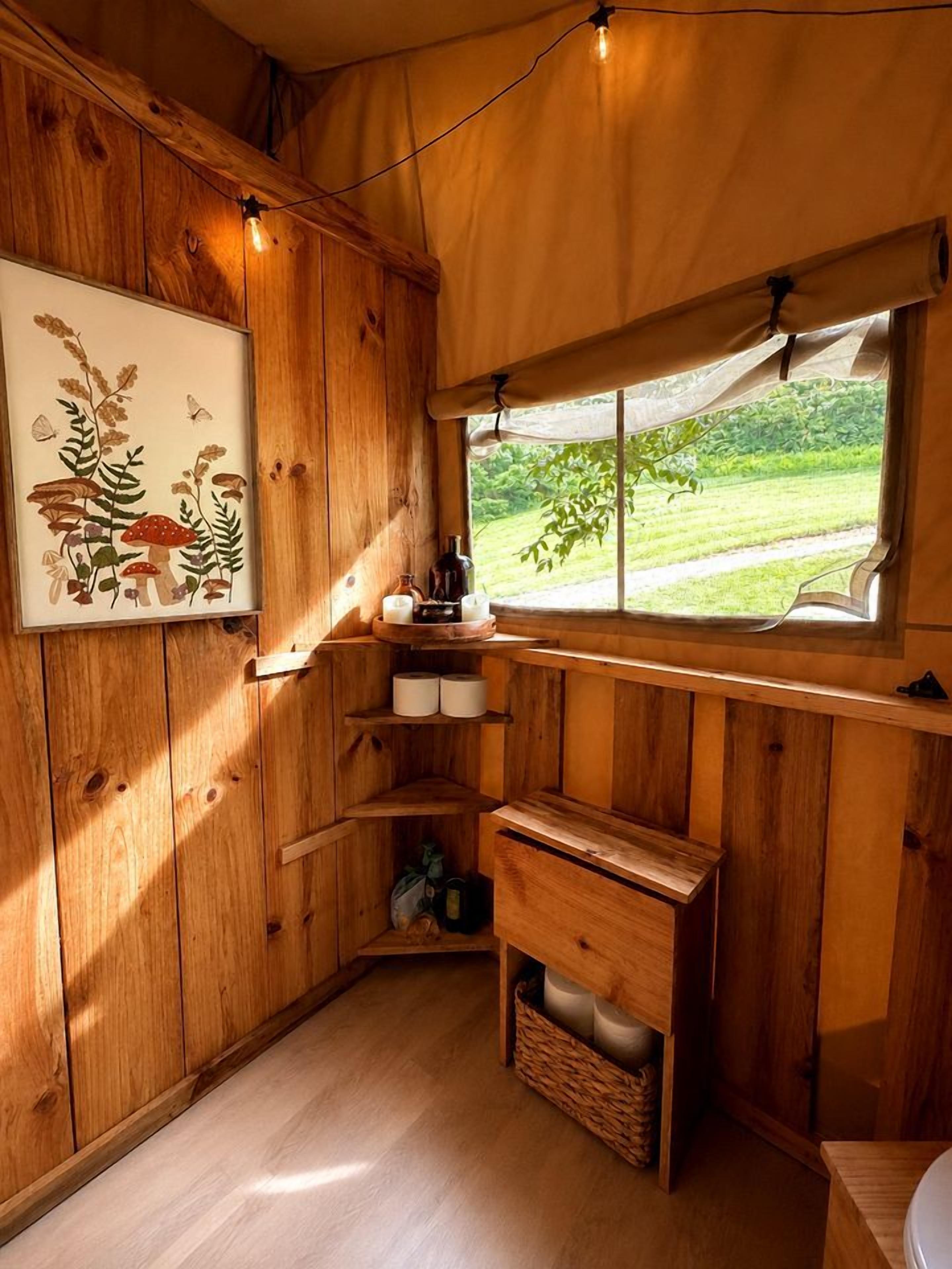 A wooden cabin interior with a window overlooking a green landscape, a shelf with candles, and a framed botanical illustration.
