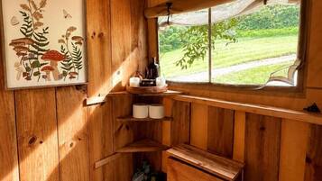 L’intérieur d’un chalet en bois, avec une fenêtre donnant sur un paysage verdoyant, une étagère avec des bougies et une illustration botanique encadrée.