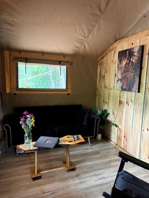 Une pièce aménagée avec une tente, un mur en bois, un canapé noir, une petite table sur laquelle est posé un vase de fleurs, un ordinateur portable et un tableau.