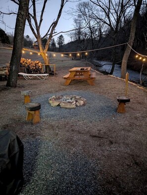A rustic outdoor setting with a wooden picnic table, a fire pit, and string lights. - Creekfront Safari Tent with Outdoor Bath and Deck in Virginia (Hillsville)