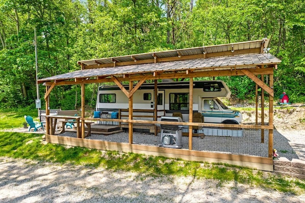 A covered RV with a picnic table and chairs, surrounded by a wooden deck and gravel.