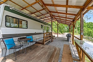 A covered RV with a patio area, including a table and chairs, a grill, and a trash can.