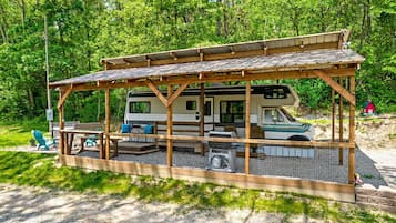 A covered RV with a picnic table and chairs, surrounded by a wooden deck and gravel.