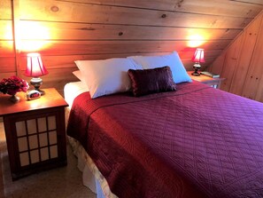 A bedroom with a wooden ceiling, a bed with a red quilt, white pillows, a dark red pillow, and two bedside lamps. - Romantic Log Cabin with Hot Tub near Cherokee National Forest, Tennessee (Coker Creek)