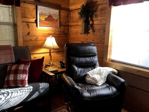 A cozy room with a leather chair, a sofa, a table with a lamp, and a window with a view. - Romantic Log Cabin with Hot Tub near Cherokee National Forest, Tennessee (Coker Creek)