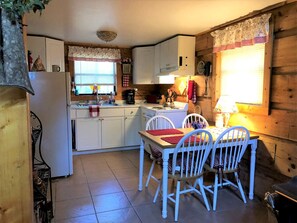 A compact kitchen with white cabinets, a dining table with chairs, and a window with blinds. - Romantic Log Cabin with Hot Tub near Cherokee National Forest, Tennessee (Coker Creek)
