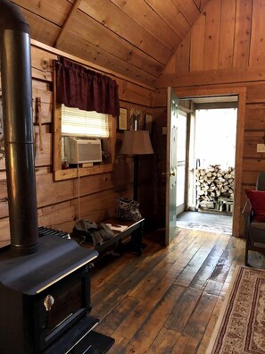 L’intérieur d’une cabane en bois, avec un poêle à bois, un banc, une lampe et une porte donnant sur l’extérieur.