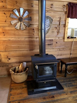 Una estufa de leña con chimenea de metal, una cesta de mimbre con leña y una pared de madera rústica.