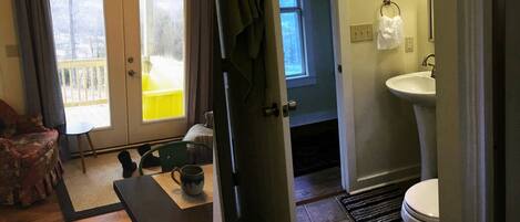 Une pièce avec un plancher en bois, une table à manger avec des chaises, un banc, une fenêtre avec des rideaux, une porte, une salle de bain avec un lavabo et des toilettes, et un petit tapis.